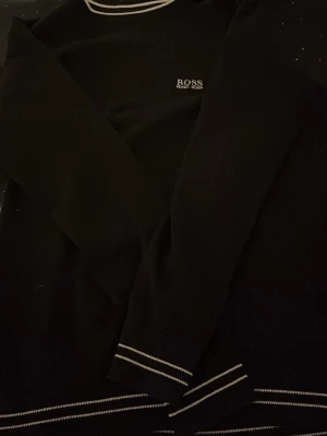 Svart tröja från BOSS med vita detaljer - Snygg svart långärmad tröja från BOSS med vita kontrastsömmar vid hals, ärmslut och nederkant. Diskret BOSS-logga på bröstet. Perfekt för en clean och stilren look.