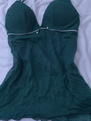 Grön spetsbaddräkt från Intimissimi - Mörkgrönt linne från Intimissimi med vadderade kupor och halterneck. Materialet är genombruten spets med små prickar och en söt detalj med vita pärlor och kantband under bysten. Perfekt för dig som vill ha en unik och feminin look