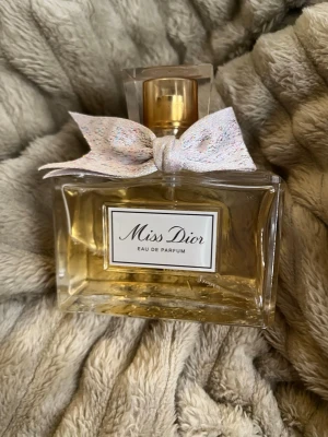 Miss Dior Eau de Parfum med rosett - Miss Dior Eau de Parfum från Dior i en elegant, rektangulär glasflaska med guldfärgad spray och en söt, ljusrosa rosett med diskreta färgdetaljer runt halsen. Flaskan har en klassisk etikett framtill och utstrålar lyxig och feminin känsla.