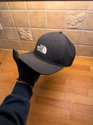 Svart keps från The North Face - Snygg svart keps från The North Face med böjd skärm och justerbart spänne bak. Klassisk design med vit broderad logga framtill. Tillverkad i slitstarkt material som passar perfekt till streetwear och casual outfits.
