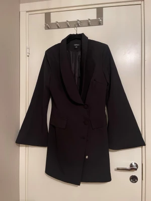 Svart kavaj med vida ärmar från NA-KD - ✨Stilren svart kavaj/kavajklänning med markerad krage och vida, långa ärmar. Kavajen har knappar framtill och fickdetaljer. Perfekt för en trendig och elegant look. Passar även en strl 38. Aldrig använd- utan prislapp✨