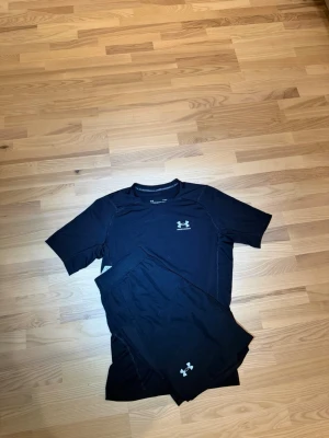 Svart träningsset från Under Armour - Säljer ett svart träningsset från Under Armour med t-shirt och shorts. Båda plaggen har Under Armour-loggan tryckt framtill. Materialet är snabbtorkande och stretchigt, perfekt för intensiva träningspass eller löpning.tröjan är L men passar S. shortsen är S 