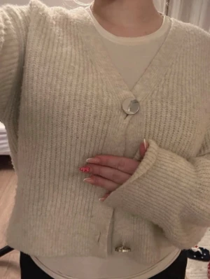 Krämvit ribbstickad kofta - Mysig krämvit kofta med ribbstickat mönster och stora dekorativa knappar framtill. Koftan har en avslappnad passform, v-ringning och långa ärmar med breda muddar. Perfekt för lager på lager och enkel att styla till olika outfits.