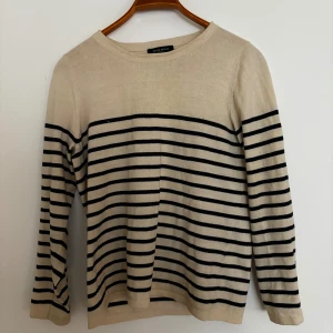 Randig stickad tröja från Bruuns Bazaar - Snygg beige stickad tröja från Bruuns Bazaar med svarta horisontella ränder. Tröjan har rund halsringning och långa ärmar. Perfekt för dig som gillar klassisk och stilren design med en modern twist.
