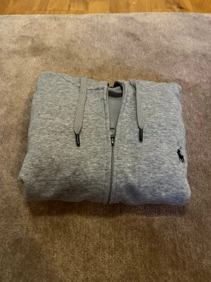 Grå zip hoodie från Polo Ralph Lauren - Snygg grå hoodie med dragkedja från Polo Ralph Lauren. Klassisk design med huva och dragsnören, ribbade muddar och liten svart logga broderad på bröstet. Tillverkad i mjukt sweatshirtmaterial som är perfekt för chill dagar.