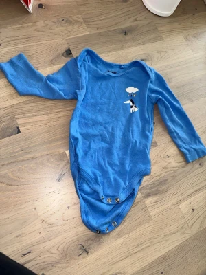 Blå långärmad body från Lupilu Lupilu blå body, storlek 74/80 - Blå body med långa ärmar och ett tryck av en hund under ett moln med regndroppar. Söt blå långärmad body från Lupilu med tryck av en hund och regnmoln på bröstet. Bodyn har praktiska tryckknappar i grenen för enkel påklädning. Tillverkad i mjuk bomull, perfekt för små barn.