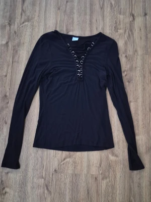 Svart långärmad topp med snörning från ginatricot - Svart långärmad topp med djup v-ringning och snörningsdetaljer framtill. Tunn och stretchig modell som sitter snyggt på kroppen. Perfekt för dig som gillar en enkel men ändå edgy stil.