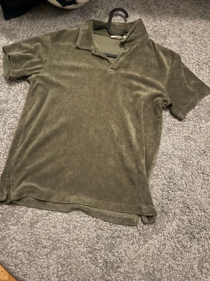 Olivgrön frottétopp med krage - Olivgrön kortärmad topp i mjuk frotté med krage och v-ringning. Perfekt för en chill och avslappnad stil. T-shirten har en loose passform och är superbekväm att bära.