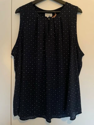 Mörkblå ärmlös blus från Holly & Whyte - Snygg mörkblå ärmlös blus från Holly & Whyte med små vita bokstäver som mönster. Blusen har rund halsringning och ett litet dekorativt veck framtill. Luftigt material som passar perfekt till jeans eller kjol.