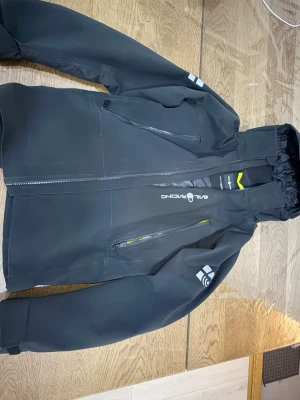 Svart vindjacka från Sail Racing - Svart vindjacka från Sail Racing med huva och två snedställda bröstfickor med dragkedja. Jackan har vita logotryck på bröstet och ärmarna samt detaljer i gult vid dragkedjorna. Tillverkad i slitstark polyester och har en sportig, modern look. XS men är lite större i storleken så passar S.