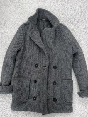 Grå cardigan från Falconeri - Stilren grå cardigan från falconeri i ribbstickad ull. Klassisk dubbelknäppning med svarta knappar och två stora fickor framtill. Hög krage och långärmad modell som passar perfekt till kyligare dagar.