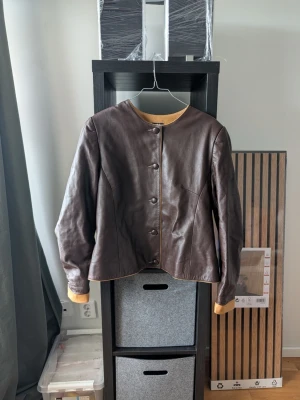 Brun skinnjacka med knappar - Snygg brun skinnjacka med rund halsringning och knäppning framtill. Jackan har långa ärmar och kontrasterande beige detaljer vid ärmslut och krage. Perfekt för dig som vill ha en stilren och klassisk look.