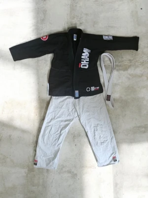 Svart BJJ gi från Manto med bälte - Säljer en svart brasiliansk jiu-jitsu gi från Manto med vita byxor och vitt bälte. Jackan har flera tryck och broderade detaljer i vitt och rött, samt förstärkta sömmar. Byxorna är vita med små loggor vid bensluten. Perfekt för träning eller tävling.