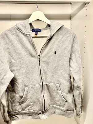 Grå zip hoodie från Polo Ralph Lauren - Klassisk grå hoodie med dragkedja från Polo Ralph Lauren. Hoodien har huva, två stora fickor framtill och den ikoniska lilla loggan broderad på bröstet. Tillverkad i mjuk bomullsblandning för skön vardagsstil.