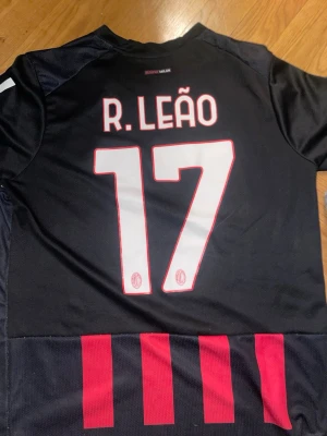 AC Milan matchtröja R. Leão 17 Puma storlek L - Säljer en svart AC Milan matchtröja från Puma med R. Leão och nummer 17 på ryggen. Tröjan har röda detaljer och ränder nedtill samt sponsortryck på ärmen. Ärmslutet har italienska flaggans färger. Perfekt för fotbollsfans! 