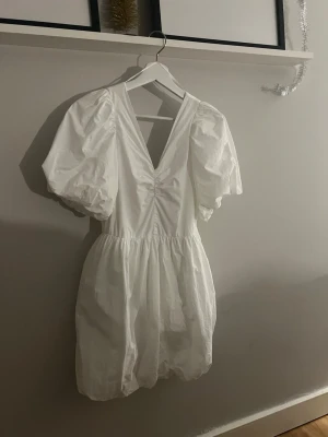 Vit kort puffärmad klänning - Supersöt vit kort klänning med puffärmar och v-ringning både fram och bak. Klänningen har en lätt rynkad midja och ballongliknande kjol. 
