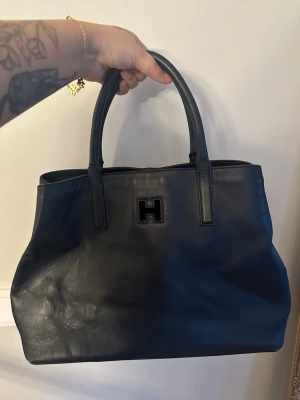 Tommy hilfiger Mörkblå handväska i skinn med H-detalj - Stilren svart handväska i skinn med dubbla handtag och en tydlig H-detalj framtill. Väskan har en klassisk, rektangulär form och rymmer det mesta du behöver till vardags. Perfekt för dig som gillar minimalistisk design och tidlösa accessoarer.