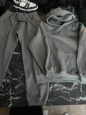 Essentials tracksuit BARA HOODIE KVAR - Säljer ett grått loungewear-set från Fear of God Essentials. Setet består av en hoodie med huva och tryck både fram och bak samt matchande mjukisbyxor med dragsko och resår i benslut. Perfekt för chill dagar hemma eller streetwear-stil.