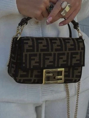 Handväska med FF-mönster - Säljer en ikonisk handväska med det klassiska FF-logomönstret i svart och brunt. Väskan har guldfärgade detaljer, kedjerem och en stor metallspänne framtill. Använd ett fåtal gånger och är i otrolig kvalitet! Dustbag tillkommer☀️