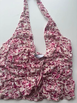Blommig halterneck topp i rosa toner - Supersöt halterneck topp med blommigt mönster i rosa och rött på ljus botten. Toppen har snörning framtill och är croppad med elastisk midja. Perfekt för varma dagar och ger en riktigt somrig vibe.