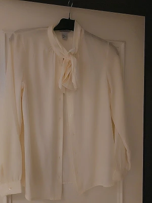 Krämvit blus med knytband - Snygg krämvit blus med knytband i halsen och knappar framtill. Blusen har långa ärmar med diskreta muddar och är tillverkad i ett lätt, flytande tyg som ger en elegant look. Perfekt att styla med jeans eller kjol för en chic outfit.