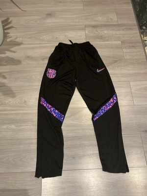 Svarta Nike FC Barcelona träningsbyxor - Säljer ett par svarta träningsbyxor från Nike med FC Barcelona-logga på ena benet. Byxorna har lila och rosa mönstrade detaljer över knäna och dragsko i midjan. Perfekta för fotbollsträning eller chill. Materialet är lätt och andas.