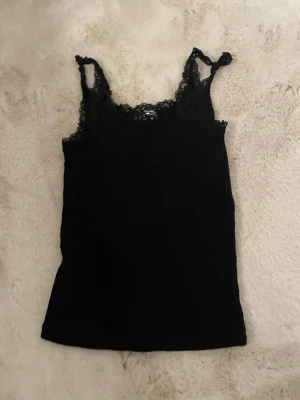 Svart spetslinnetopp från Divided - Säljer en svart linnetopp från Divided med fina spetsdetaljer vid axelbanden och halsringningen. Toppen är ärmlös och har en slim passform som sitter snyggt på kroppen. Perfekt att styla med jeans eller kjol. Dock knuten i axelbanden därav priset.