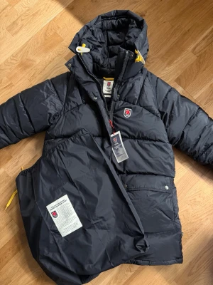 Svart dunjacka Fjällräven Expedition - Svart dunjacka från Fjällräven Expedition Series med gul dragsko och klassisk logga på bröstet. Jackan har huva, stora fickor och quiltad design. Perfekt för kalla vinterdagar och riktigt varm. 