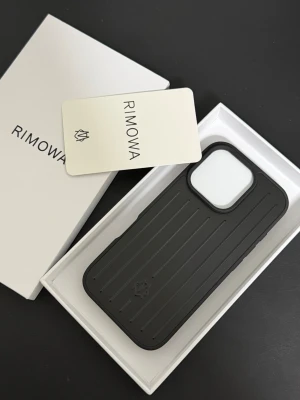 Svart iPhone 16 pro Rimowa mobilskal - Snyggt svart mobilskal från Rimowa med ikoniska räfflade linjer och diskret logga. Skalet är gjort i tåligt material och har en robust design som skyddar din iPhone. Perfekt för dig som vill ha en stilren och exklusiv look på mobilen.