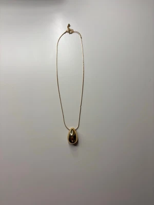 Guldigt halsband med droppformad berlock - Snyggt halsband med tunn guldfärgad kedja och en stor, droppformad berlock i metall. Från glitter