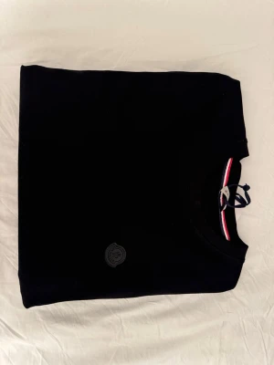 Svart t-shirt från Moncler - Stilren svart t-shirt från Moncler med rund halsringning och diskret Moncler-logga på bröstet. Klassisk passform och snygg detalj med röd, vit och blå rand på insidan av kragen. Aldrig använt.