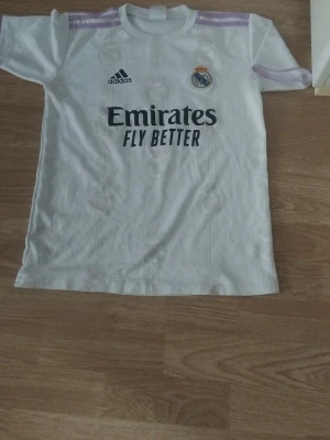 Vit Real Madrid t-shirt Adidas - Säljer en vit Real Madrid t-shirt från Adidas med klubbmärke och sponsortryck på bröstet. Tröjan har korta ärmar och lila detaljer på axlarna. Perfekt för dig som älskar fotboll och vill representera ditt favoritlag.