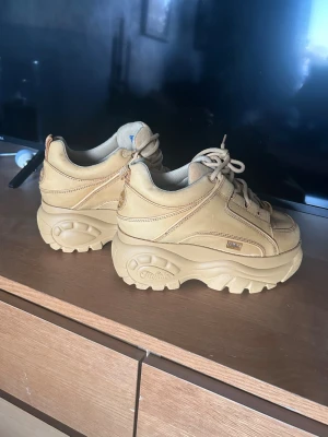 Beige Buffalo sneakers med platåsula - Buffalo sneakers i beige. Skorna har snörning och är tillverkade i syntetmaterial med tydliga Buffalo-loggor på sidan och under sulan. Har aldrig använt skorna då de va för stora för mig storlek 39. 