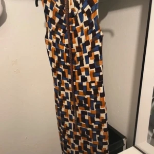 Mönstrad långklänning i retrostil - Säljer en långklänning med coolt geometriskt mönster i blått, brunt, orange och vitt. Klänningen har en figurnära passform och knytning upptill. Perfekt för dig som gillar unika och färgstarka plagg!