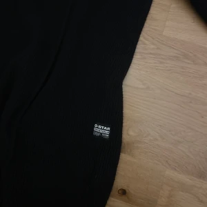 Svart stickad tröja från G-Star  - Svart stickad tröja från G-Star RAW i storlek M. Tröjan har en ribbad struktur, rund halsringning och långärmade ärmar. Diskret logotyp på sidan ger en clean och stilren look. Perfekt för dig som gillar enkel och snygg design.