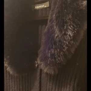 Svart kofta med päls från Jasmine - Säljer en svart ribbad kofta från Jasmine med mjuk päls krage, som går att ta bort. Koftan har långa ärmar och knappar.