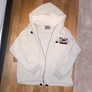 Beige hoodie från Belmondo Sports - Beige hoodie från Belmondo Sports med broderad logga på bröstet. Tröjan har dragkedja, huva med snörning och ribbade muddar. Dragkedjan är sönder och tröjan har en missfärgning på framsidan (som dock skulle kunna tvättas bort).