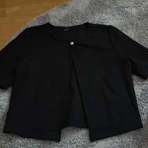 En svart kofta från Gina Tricot - Stilren svart kofta från Gina Tricot i storlek S. Jackan har korta ärmar, rund halsringning och en dekorativ guldfärgad knapp framtill. Passformen är normal och materialet känns mjukt och bekvämt. Perfekt för att addera en clean touch till din outfit.