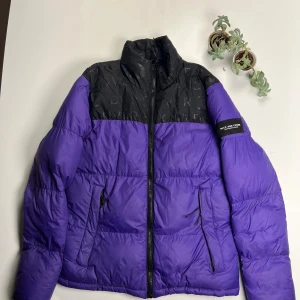 Lila och svart pufferjacka Duck and Cover - Snygg pufferjacka från Duck and Cover i lila och svart med logga på ärmen. Jackan har hög krage, dragkedja framtill och två fickor med dragkedja. Överdel i svart med diskret logomönster och resten i lila. Perfekt för kalla dagar som närmar sig snabbt! 