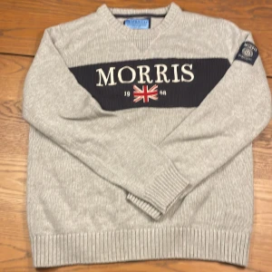 Grå stickad tröja från Morris - Snygg grå stickad tröja från Morris med broderad logga och brittisk flagga framtill. Tröjan har ribbade muddar, lång ärm och en mörkblå panel över bröstet. Perfekt för dig som gillar klassisk och sportig stil.