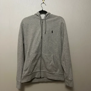 Grå zip hoodie Polo Ralph Lauren - Snygg och klassisk grå hoodie från Polo Ralph Lauren med dragkedja framtill och logo på bröstet. Väldigt skön och i bra skick! Endast ett mycket litet hål vid höger ficka (knappt synligt, se bild). Nypris ca 1895kr på Zalando.