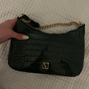 Victoria Secret väska/purse i grön färg original - Snygg grön axelväska från Victoria Secret med krokopräglat mönster och guldfärgad kedja som axelrem. Väskan har en dragkedja upptill och en elegant guldfärgad logga framtill. Perfekt för dig som vill sticka ut med en trendig accessoar.