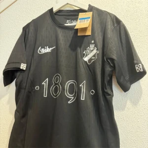 Svart AIK fotbollströja Nike M - AIK fotbollströja från Nike i svart med vita detaljer och klubbmärke på bröstet. Snyggt tryck med '1891' framtill och diskreta mönster i tyget. Tillverkad i lätt och funktionellt material, perfekt för match eller träning.