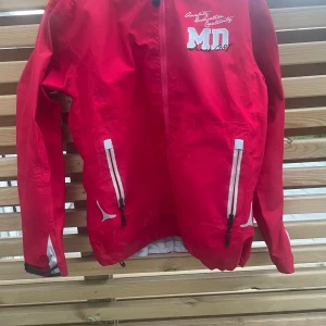 Röd vindjacka från Matterhorn, strl 38 - Röd vindjacka från Matterhorn med huva och vita detaljer. Jackan har dragkedjor framtill, broderad text på bröstet och tryck på ryggen. Perfekt för blåsiga dagar och har en sportig look med snygga kontrastdetaljer.