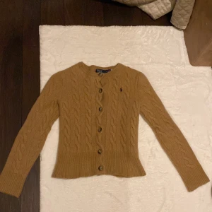 Beige kabelstickad kofta Polo Ralph Lauren - Snygg beige kabelstickad kofta från Polo Ralph Lauren med rund hals och knappar framtill. Tröjan har lång ärm, ribbad nederkant och är tillverkad i en lyxig mix av ull och kashmir. Diskret broderad logga i mörkblått på bröstet. Perfekt för dig som gillar klassisk stil. Krympt i samband med tvätt…. Passar som xxs dam. Skriv för mått