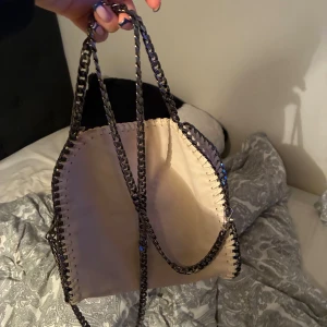 Beige axelväska Stella McCartney - Snygg beige axelväska från Stella McCartney med kedjehandtag och dekorativa sömmar längs kanterna. Väskan har dragkedja på baksidan och ett stort huvudfack. Tillverkad i syntetmaterial och har en rund metallbricka med logga.
