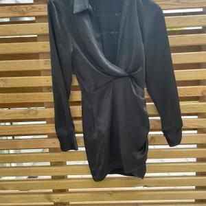 Svart omlottklänning med krage - Snygg svart kort omlottklänning med lång ärm och klassisk krage. Klänningen har en djup v-ringning och är tillverkad i ett glansigt, slätt material som ger en lyxig känsla. Perfekt för dig som gillar stilrena och trendiga plagg.