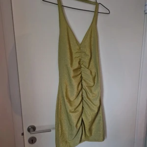 Glittrig grönl kortklänning med rynk - Supersnygg guldigt glittrig grön kortklänning med v-ringning och rynk framåt. Jag är 168 och den når till mina nedre lår. Storlek M och den är stretchig. Köpt second hand men jag har endast provat den och den är i fint skick. Katt finns i hemmet