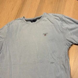 Ljusblå GANT t-shirt i bomull - Ljusblå t-shirt från GANT med klassisk rund hals och liten broderad logga på bröstet. Tillverkad i mjuk bomull och har en enkel, stilren design. Perfekt för en avslappnad look. Har några små blekta fläckar se bild man tänker bara på dem om man tittar noga 