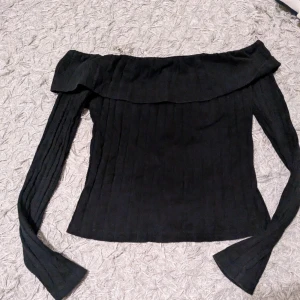 Svart offshoulder ribbad topp - Snygg svart offshoulder topp med långa ärmar och ribbad struktur. Toppen har en bred volangkant längs axlarna som ger en trendig look. Perfekt att styla med jeans eller kjol för en cool outfit.